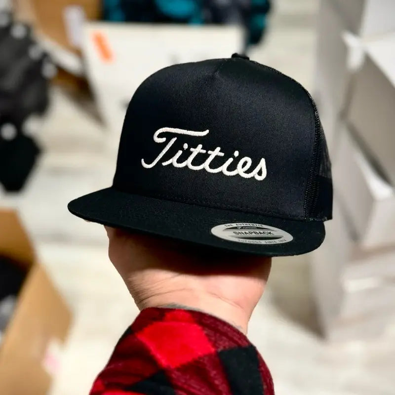 Black cap with 'T*tties' text held by a person wearing a red and black checkered shirt.