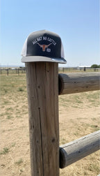 Charcoal and white trucker snapback hat with 'All Hat No Cattle' text on a wooden post with a rural landscape background.