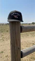 Black trucker snapback hat with 'All Hat No Cattle' text on a wooden post with a rural landscape background.