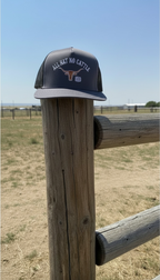 Charcoal and black trucker snapback hat with 'All Hat No Cattle' text on a wooden post with a rural landscape background.