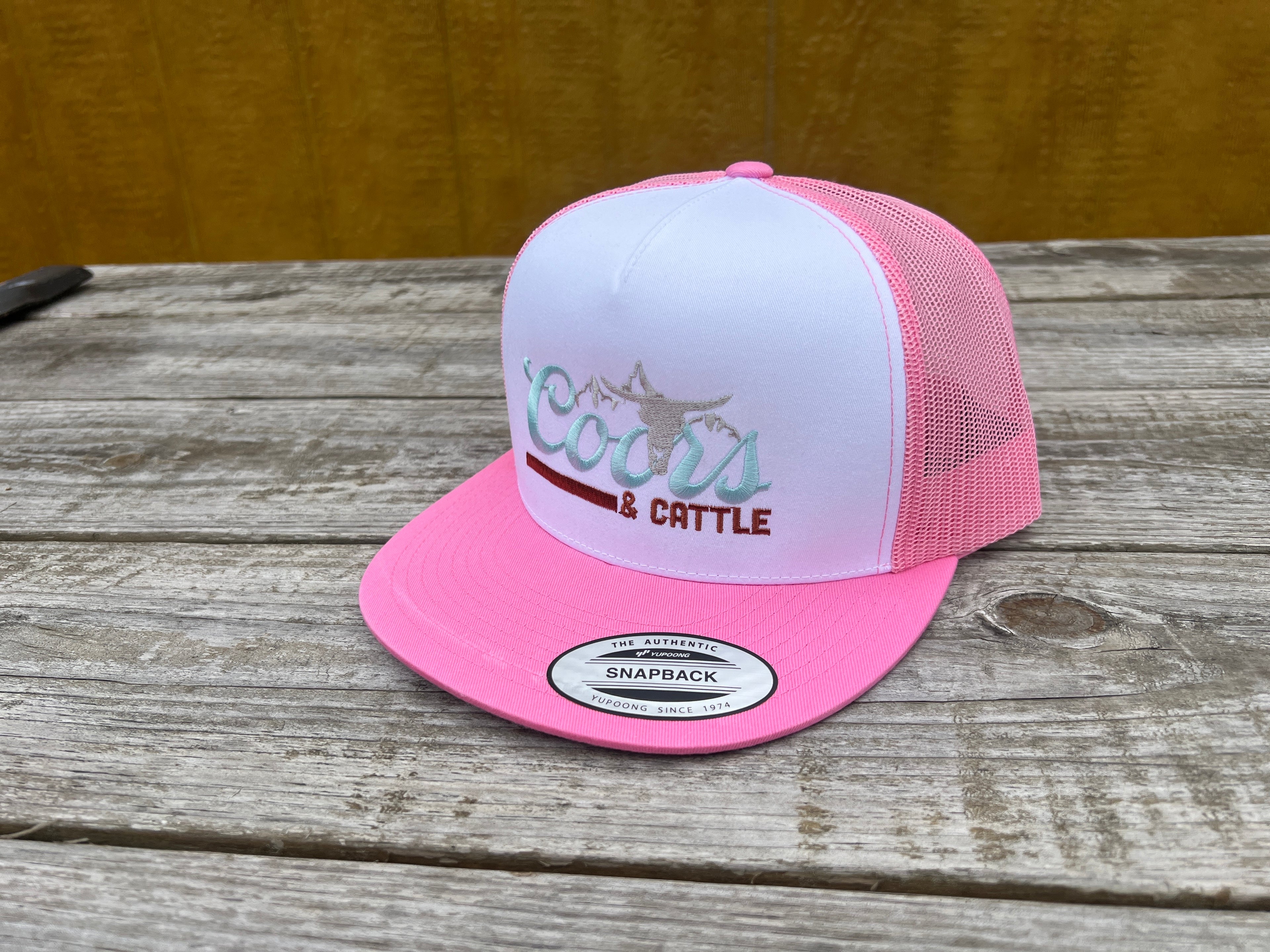 Pink and white trucker hat with 'Coors & Cattle' text on a wooden surface