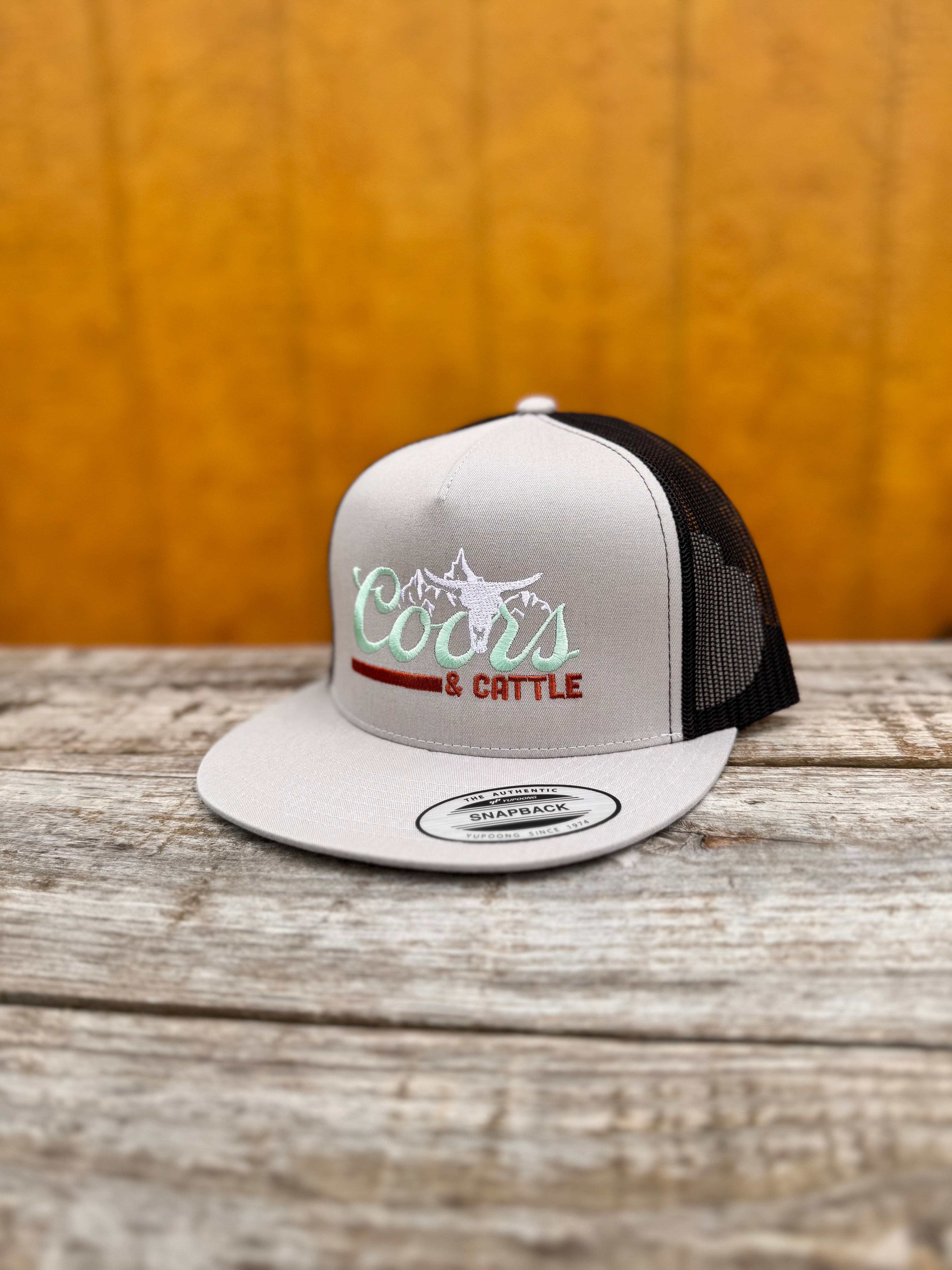 gray and black trucker hat with 'Coors & Cattle' text on a wooden surface