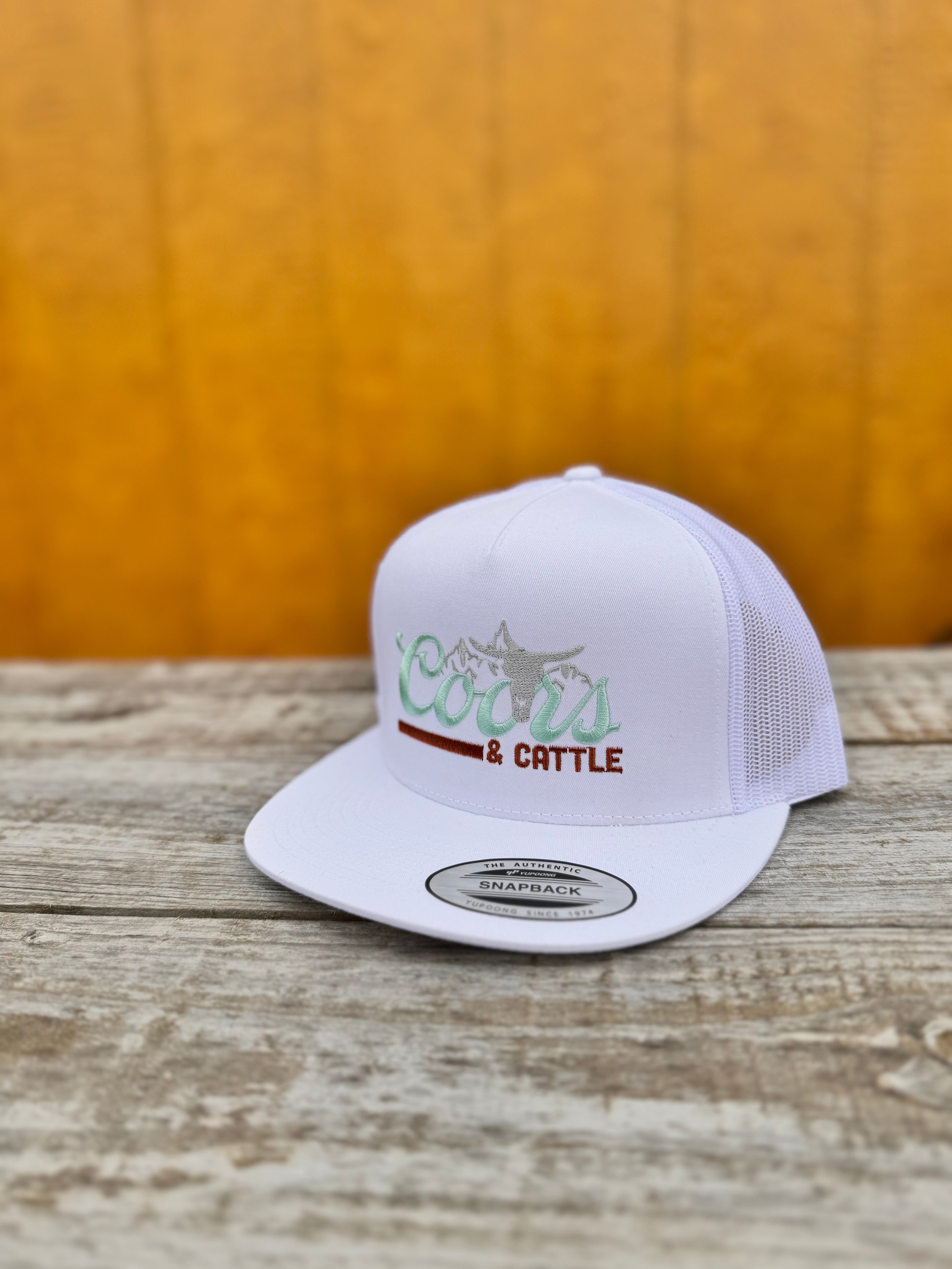 White trucker hat with 'Coors & Cattle' text on a wooden surface
