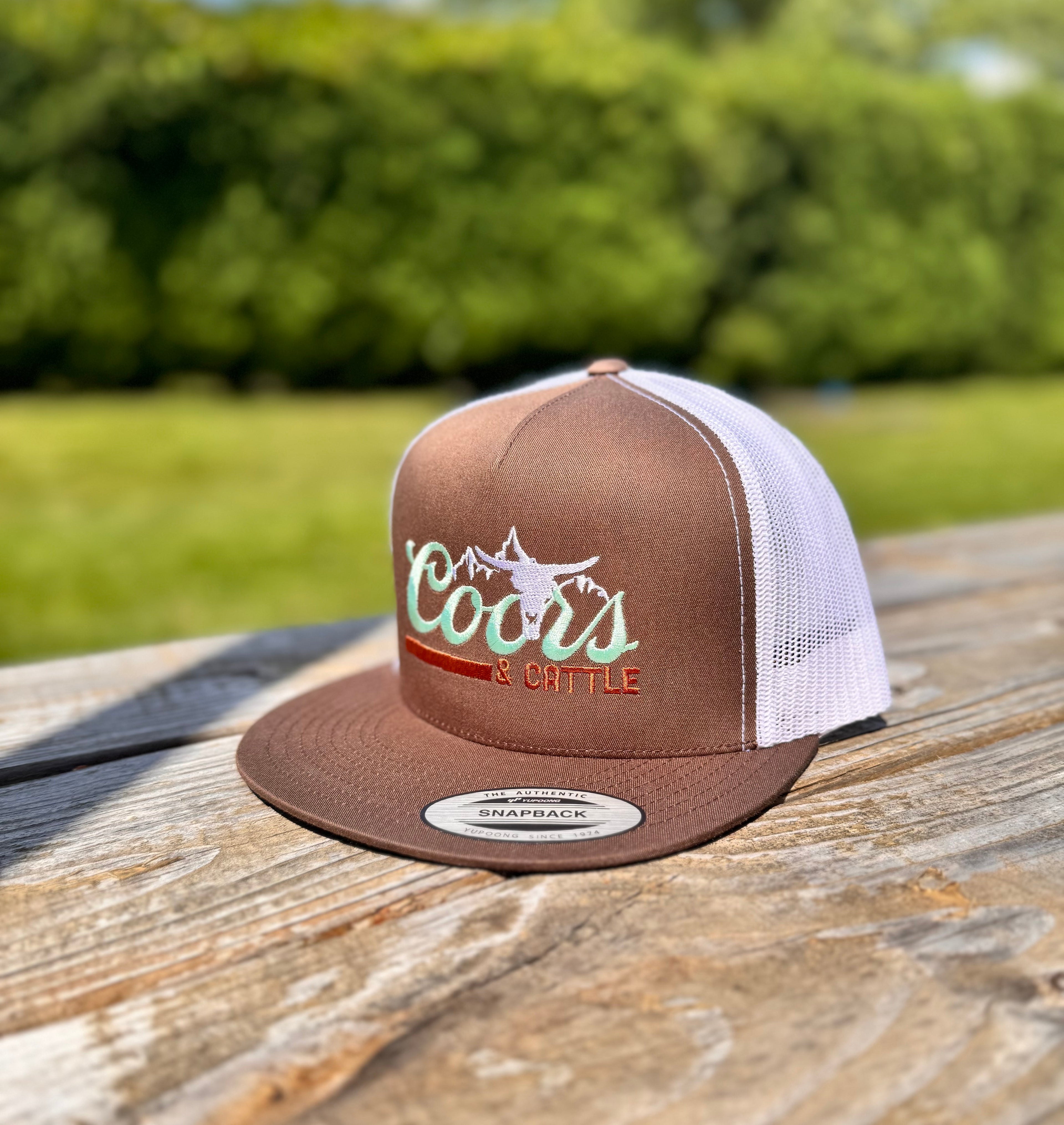 Brown and white cap with 'Coors & cattle' text on a wooden surface with blurred greenery in the background