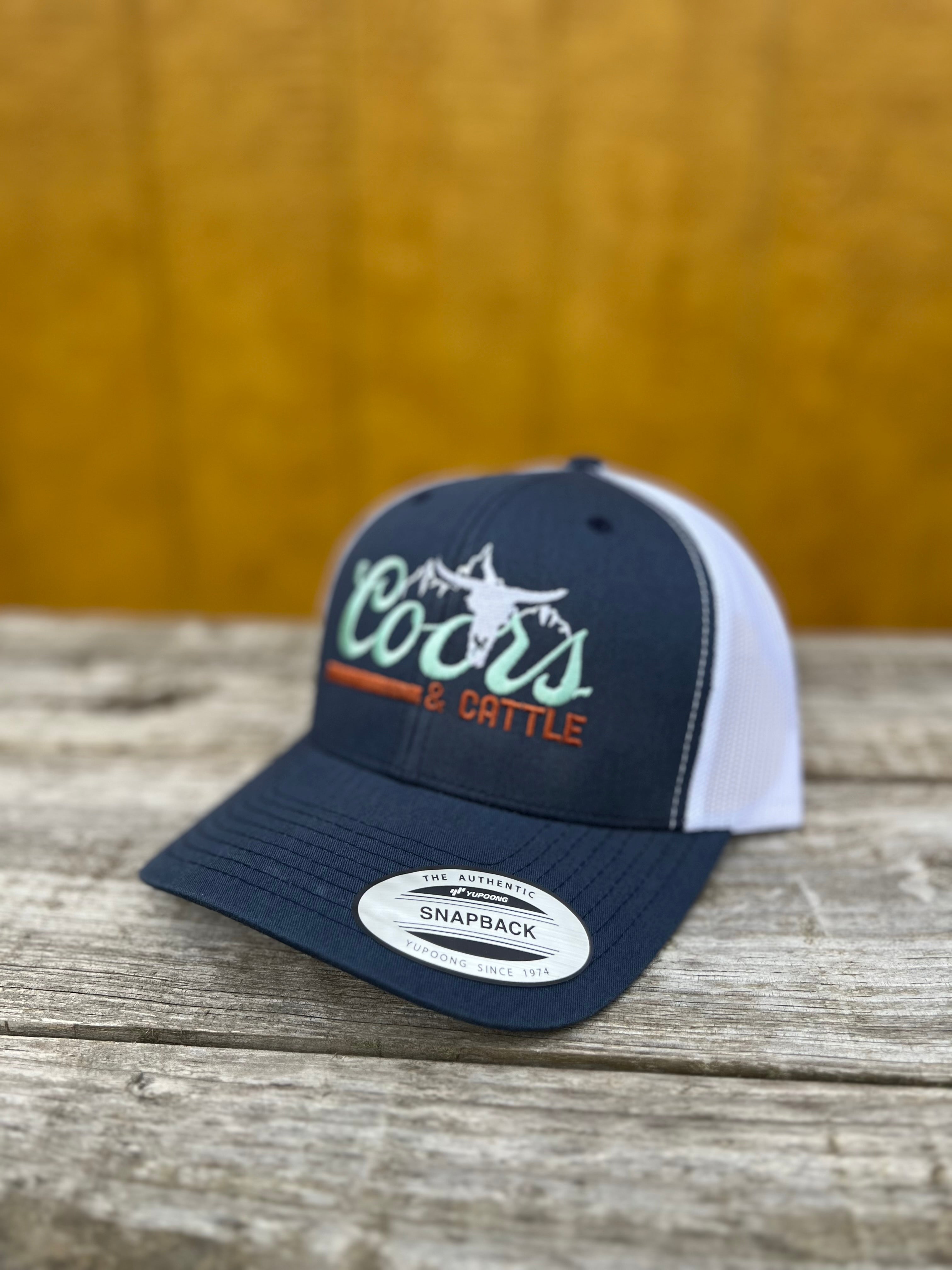 navy and white curved snapback hat with 'Coors & Cattle' text on a wooden surface