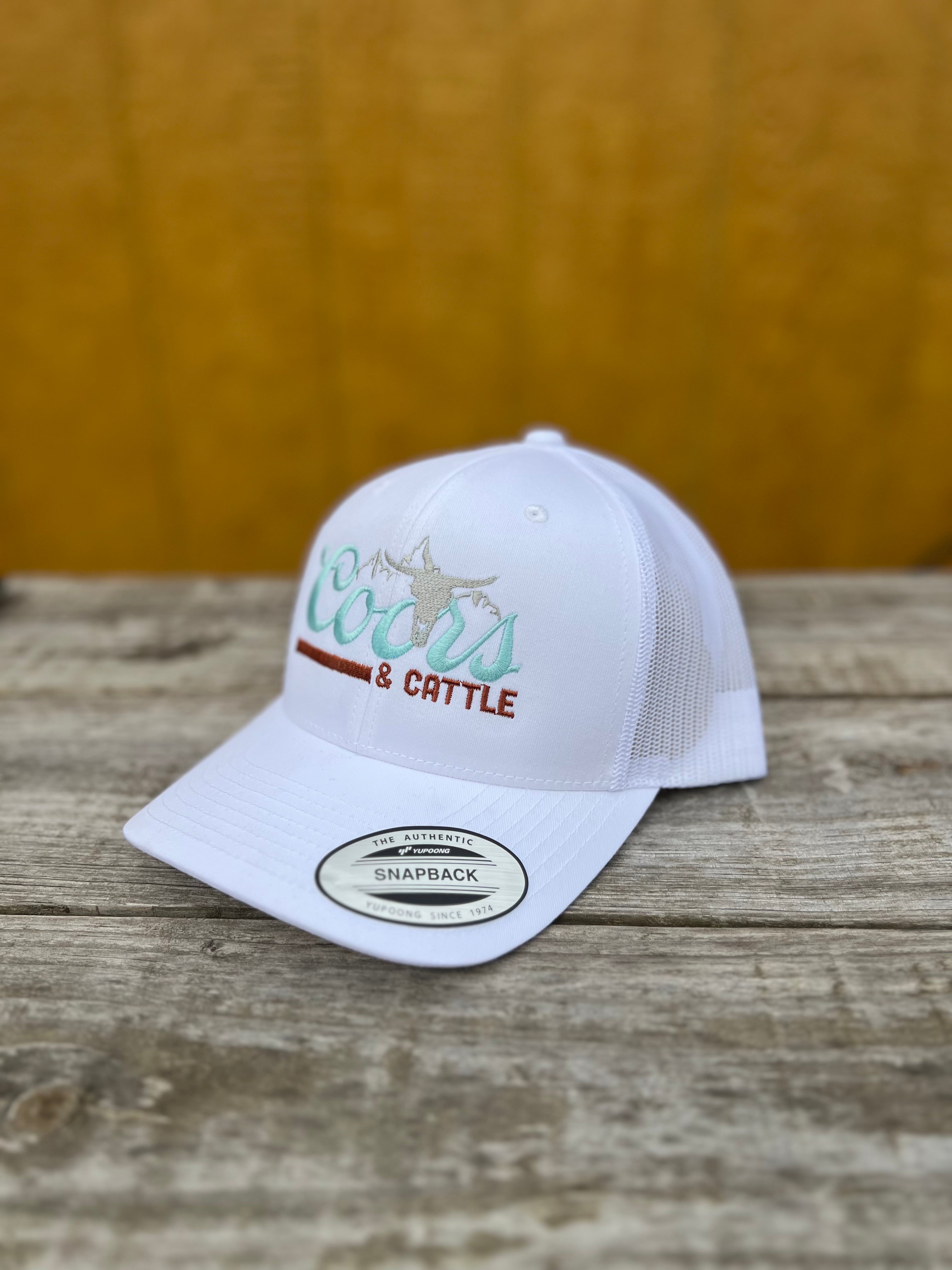 White snapback hat with 'Coors & Cattle' text on a wooden surface
