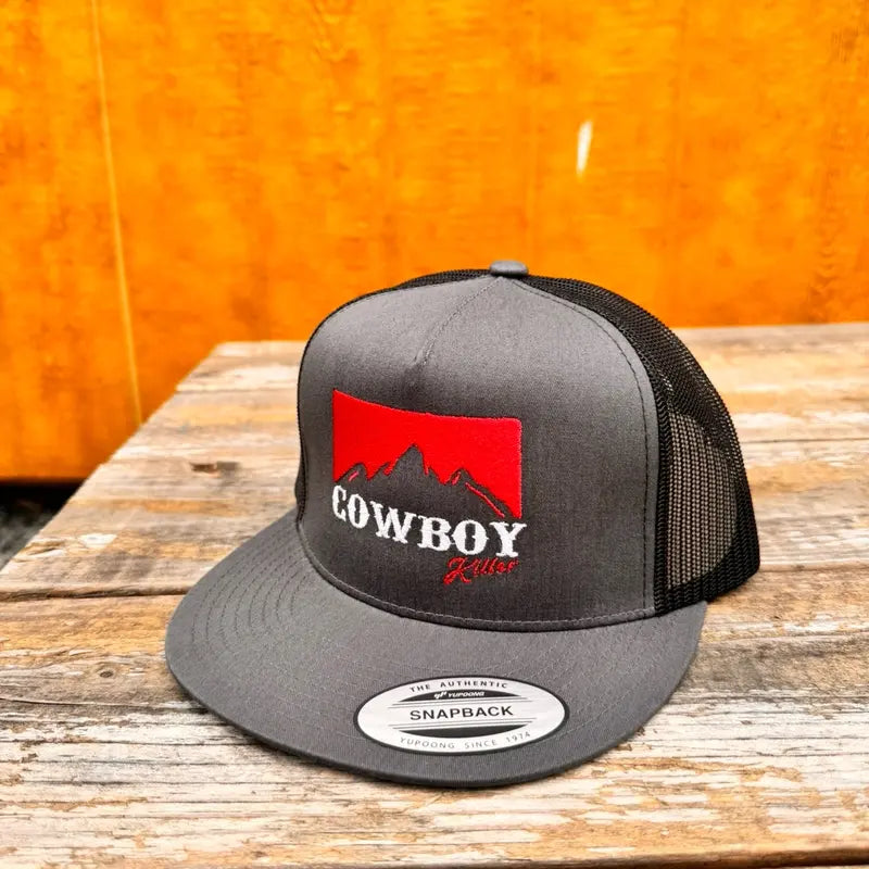 Charcoal and black trucker snapback hat with red and white 'cowboy killer' text and mountain logo on a wooden surface