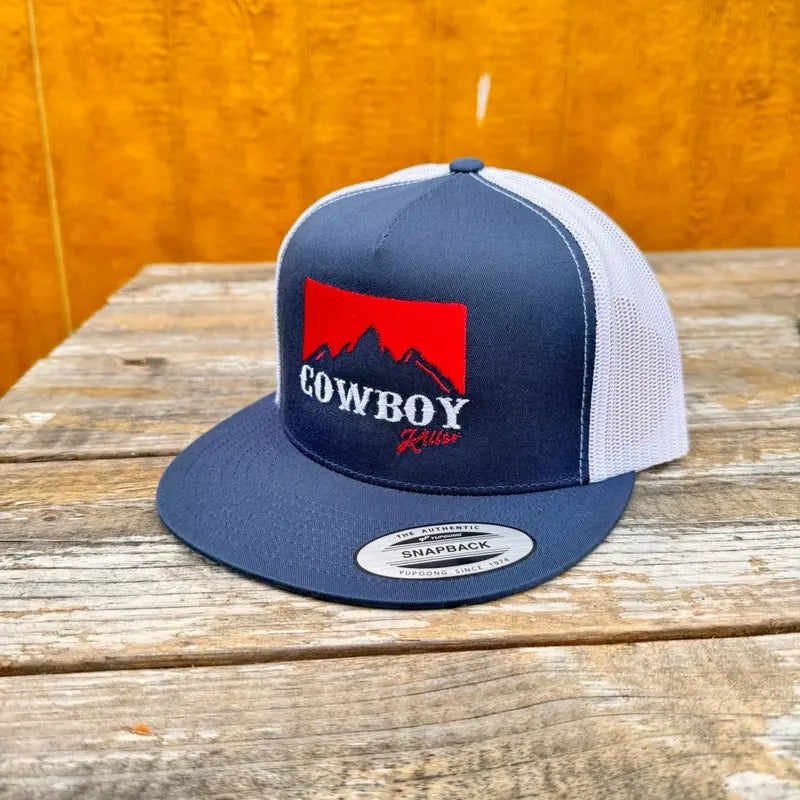 Blue and white trucker snapback hat with red and white 'cowboy killer' text and mountain logo on a wooden surface