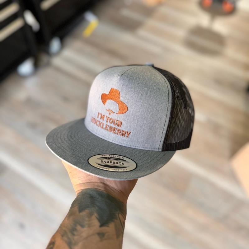 Gray and black snapback hat with orange 'i'm your huckleberry' text held by a person with tattoos.