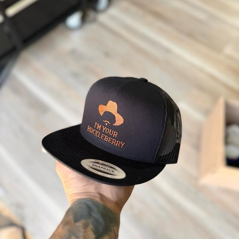 Black snapback hat with orange 'I'm Your Huckleberry' text held by a person with tattoos.