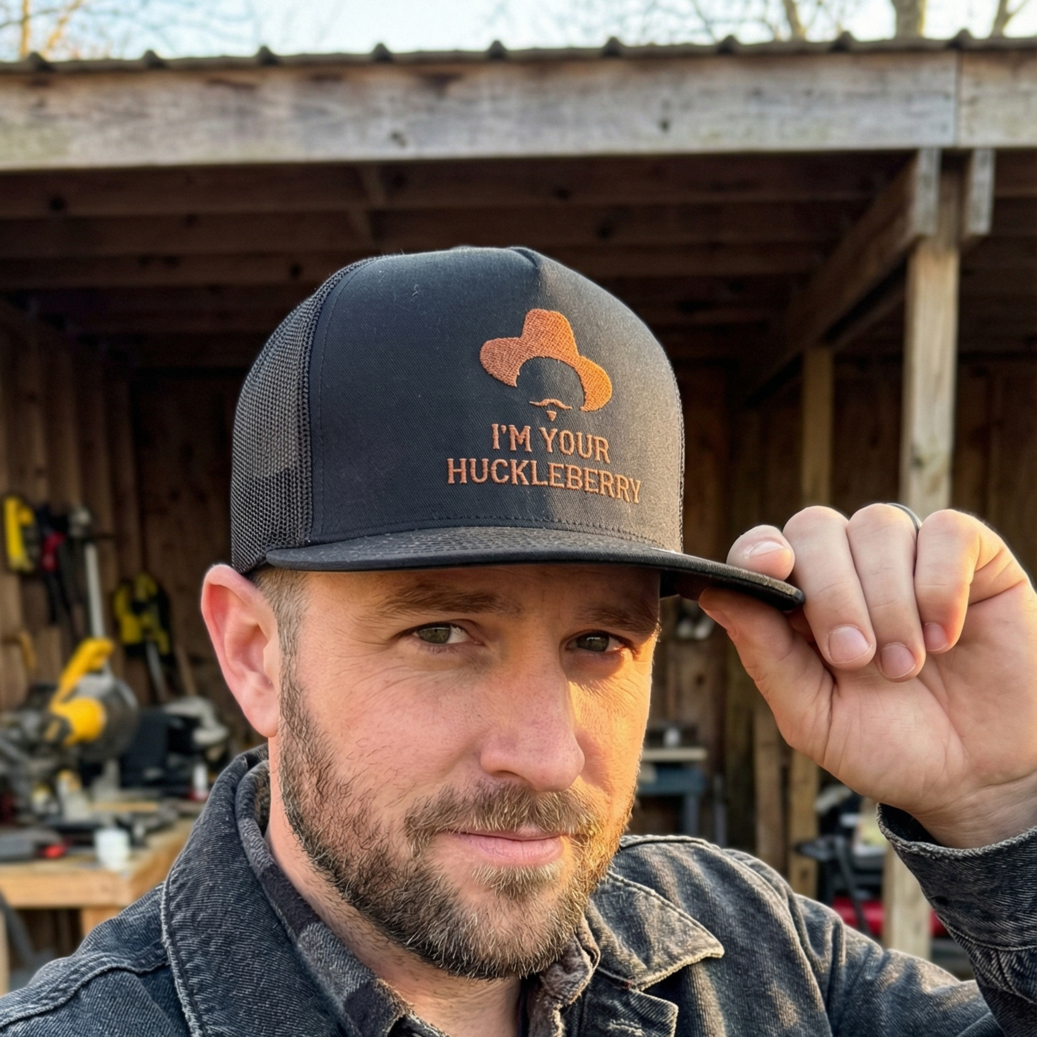 Man wearing a snapback hat with 'I'm Your Huckleberry' text in an outdoor work setting.