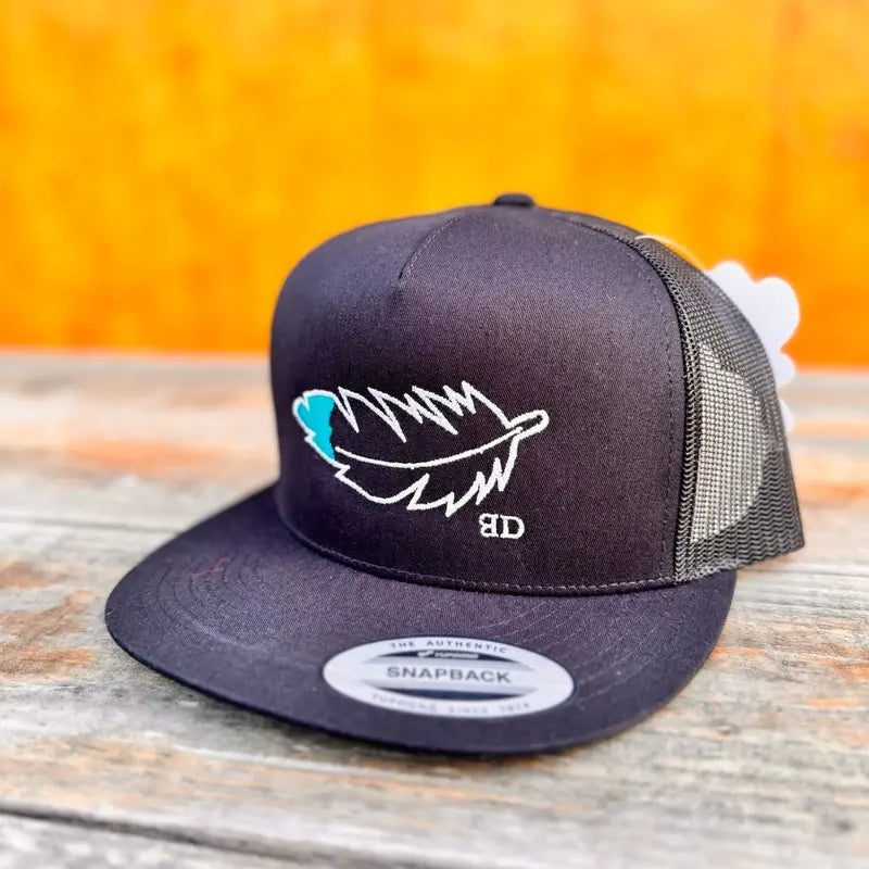 Black snapback cap with teal and white feather design on a wooden surface