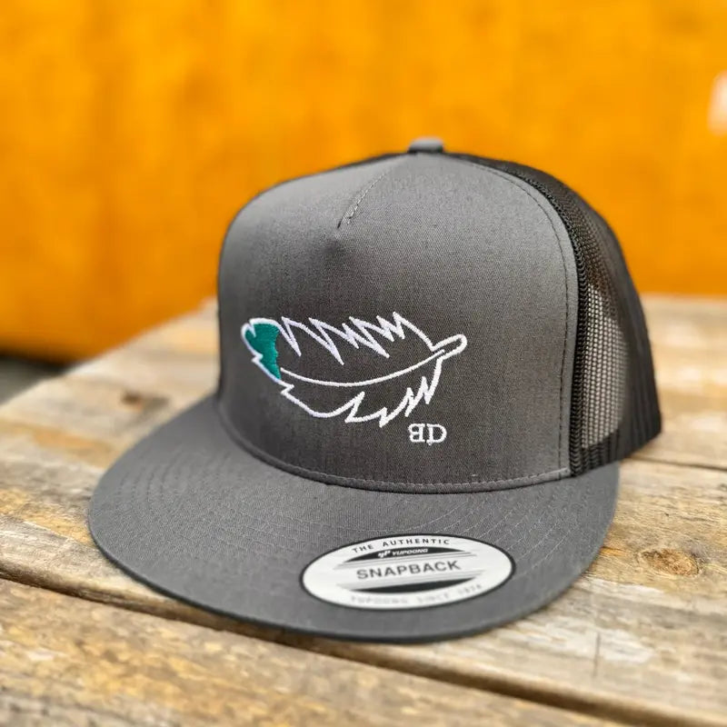 charcoal and black trucker cap with teal and white feather design on a wooden surface