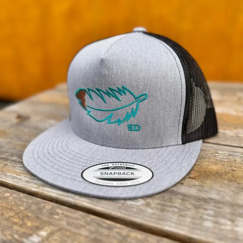 Grey and black snapback cap with teal feather design on a wooden surface