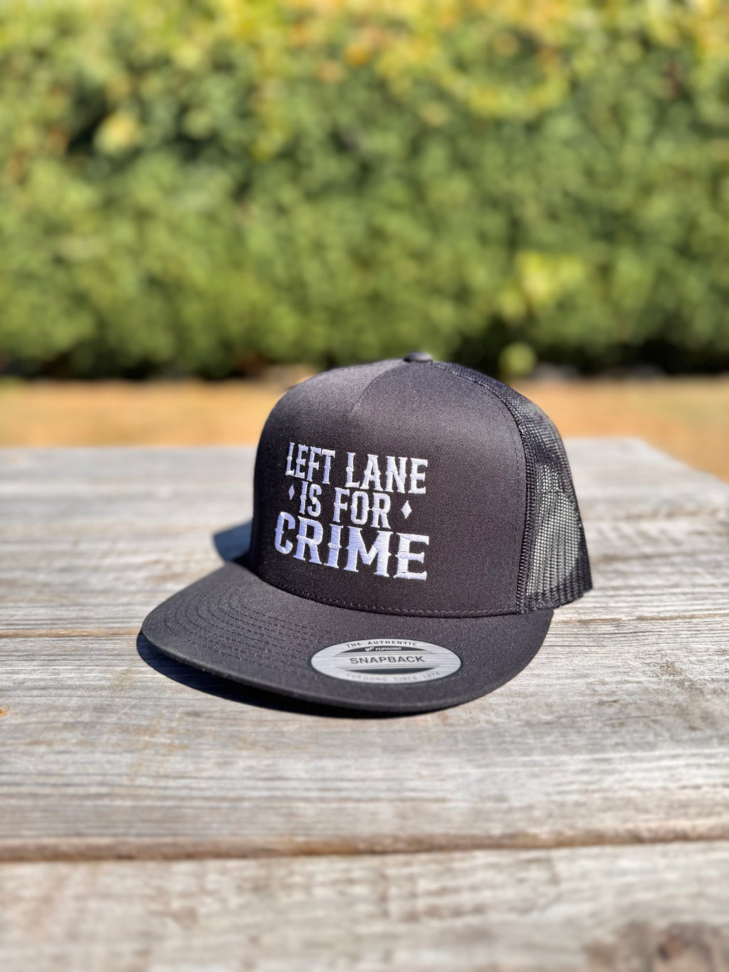 Black trucker hat with 'LEFT LANE IS FOR CRIME' text on a wooden surface with greenery in the background