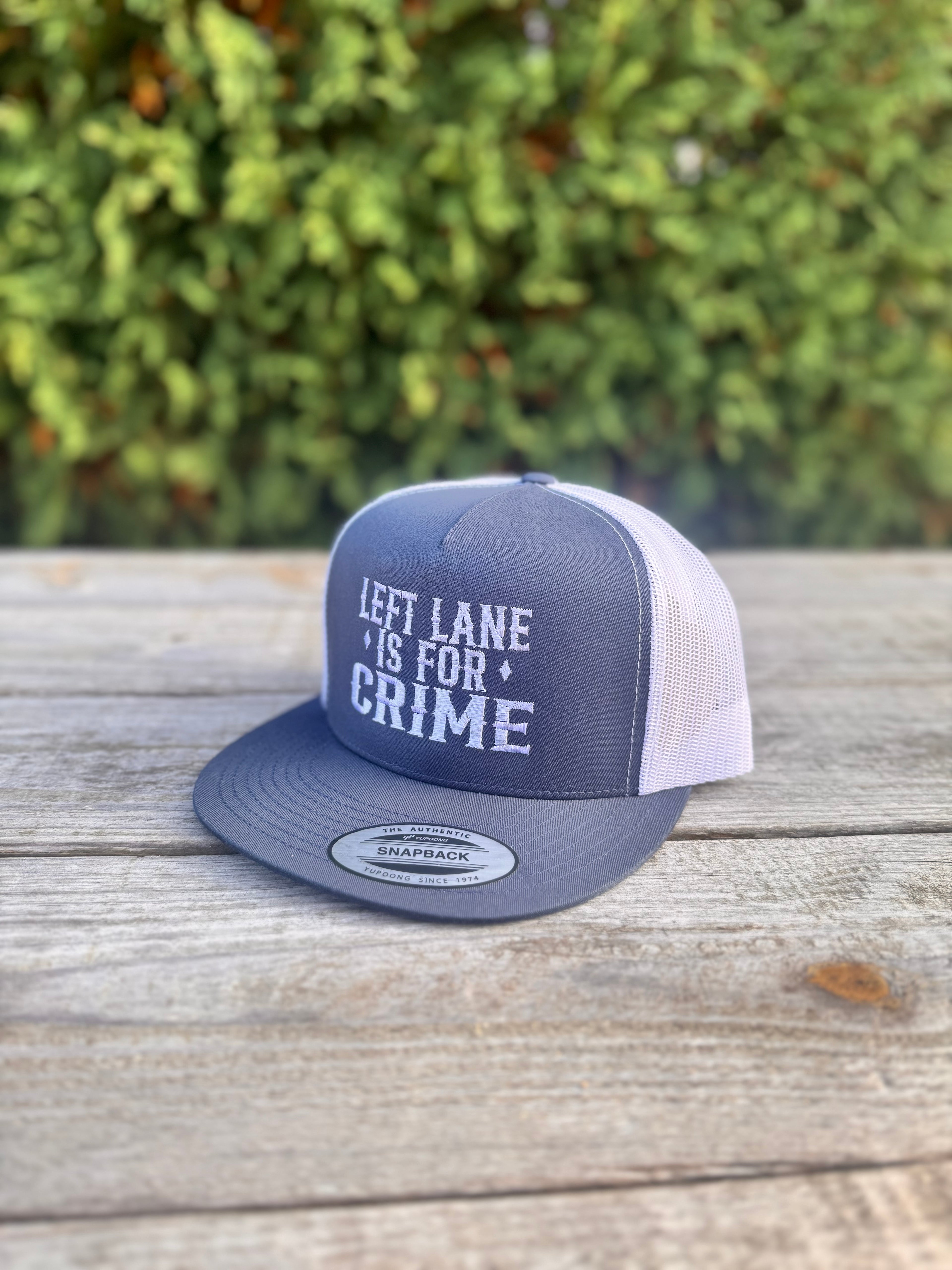 Gray and white cap with 'Left Lane is for Crime' text on a wooden surface with greenery in the background
