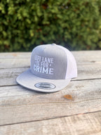 Light Gray and white cap with 'Left Lane is for Crime' text on a wooden surface with greenery in the background