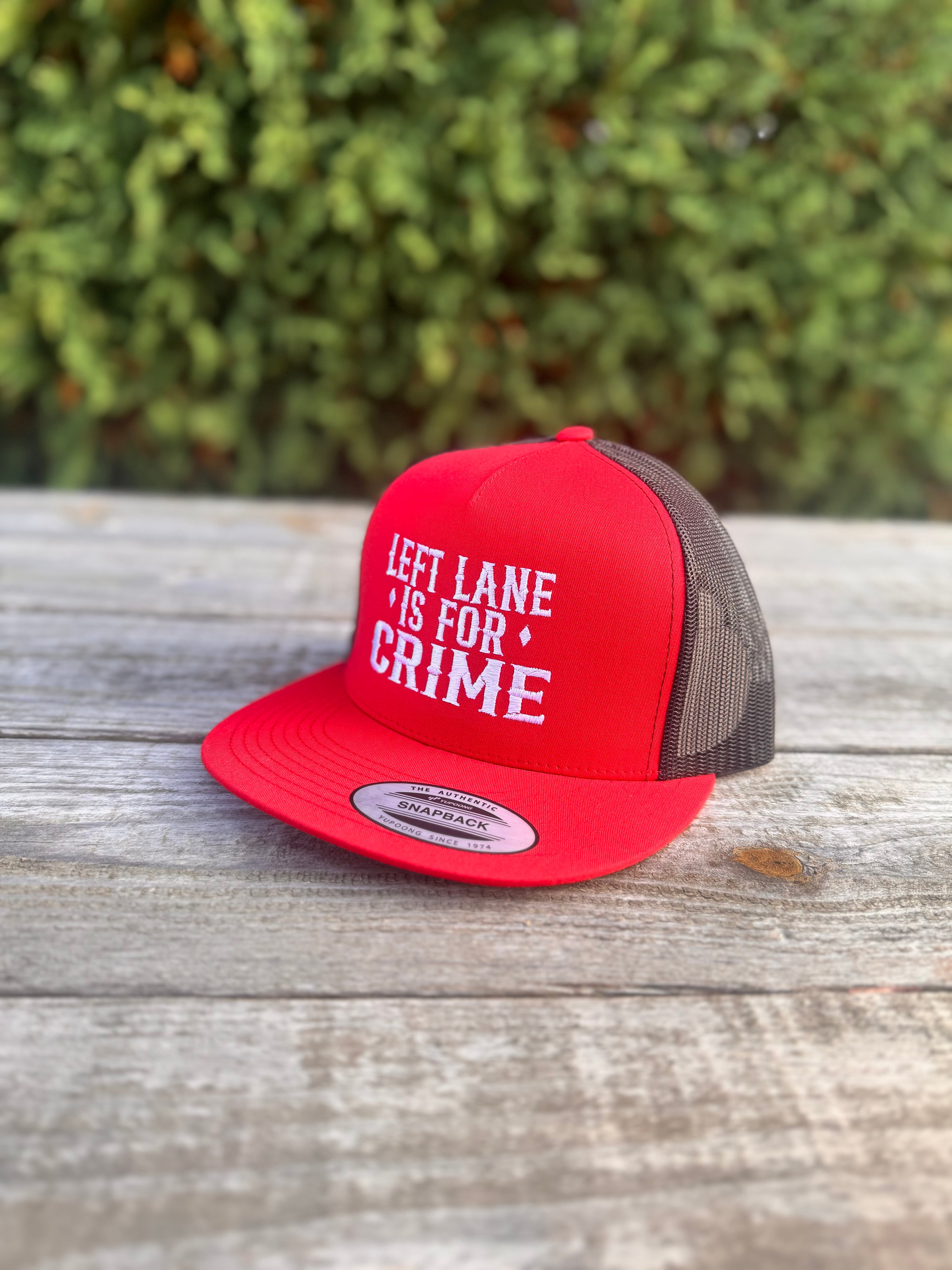 Red and black cap with 'Left Lane Is For Crime' text on a wooden surface with greenery in the background