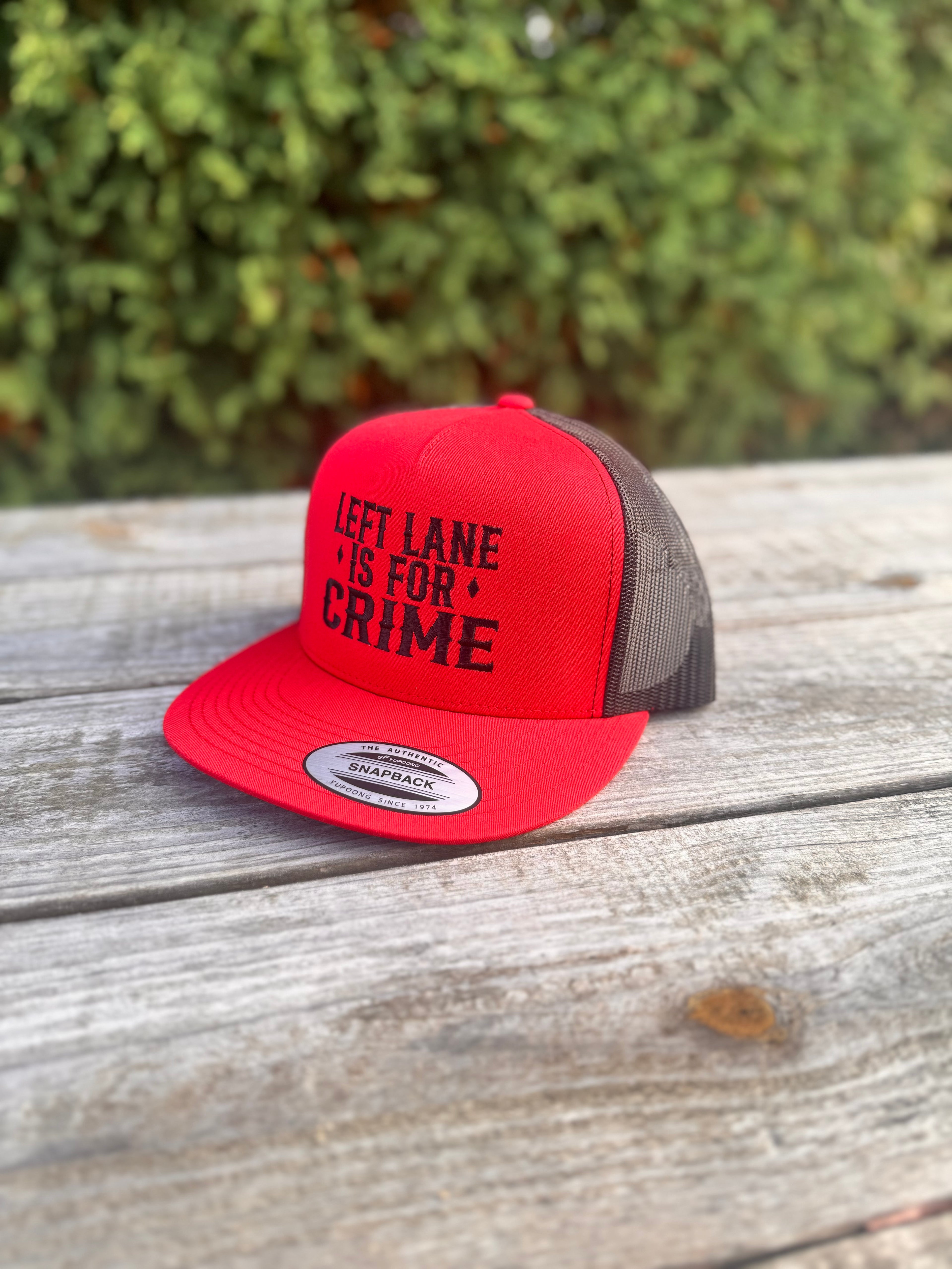 Red and black text trucker hat with 'LEFT LANE IS FOR CRIME' text on a wooden surface with greenery in the background.