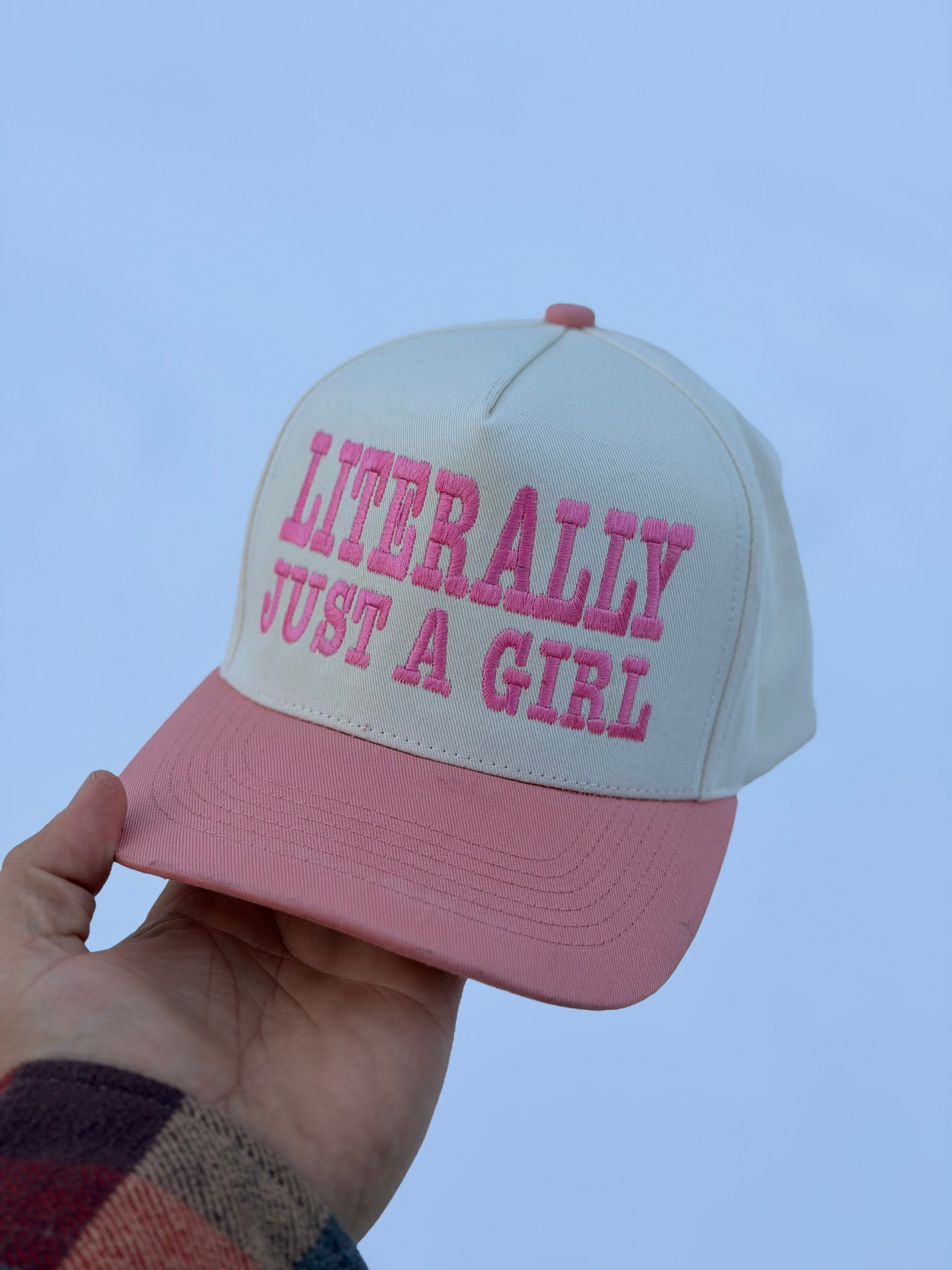 Pink and white cap with 'LITERALLY JUST A GIRL' text held against a blue background.