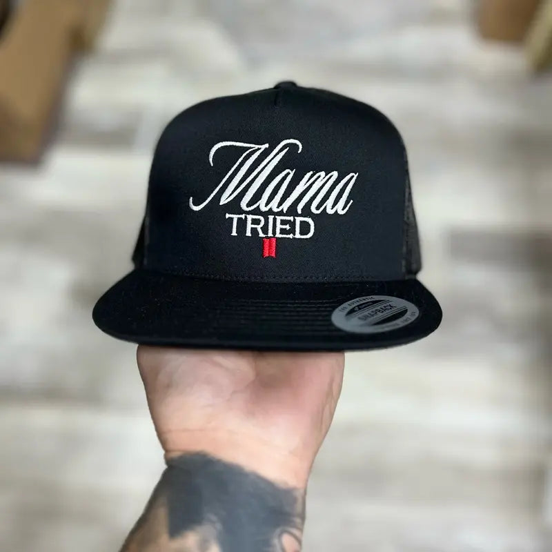 Black snapback hat with 'Mama Tried' text held by a person against a neutral background