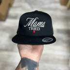 Black snapback hat with 'Mama Tried' text held by a person against a neutral background