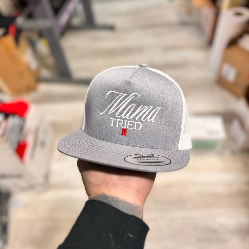 Silver and white snapback hat with 'Mama Tried' text held by a person against a neutral background