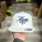 White snapback hat with 'Mama Tried' text held by a person against a neutral background