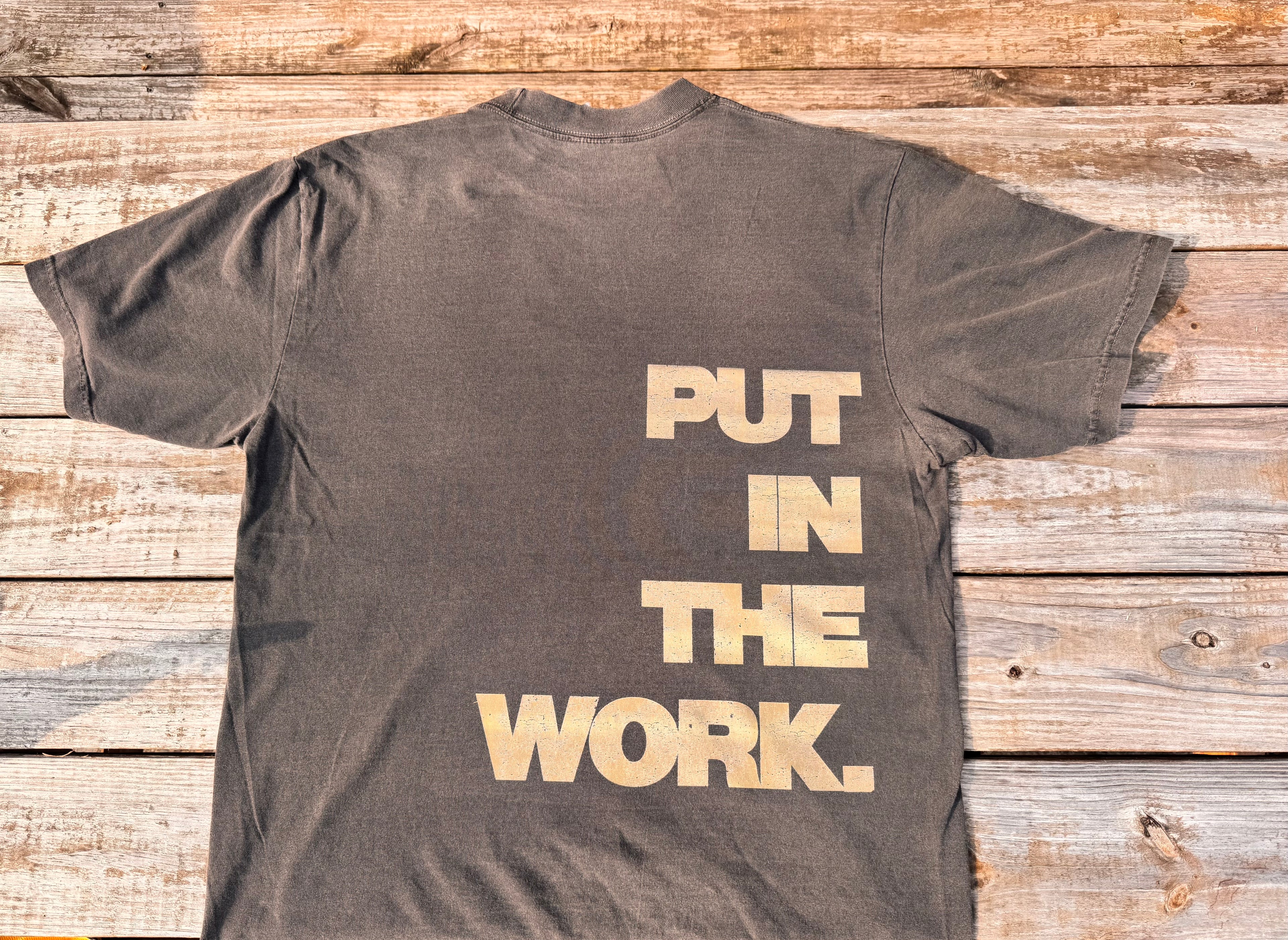 gray t-shirt with tan 'PUT IN THE WORK' text on a wooden surface