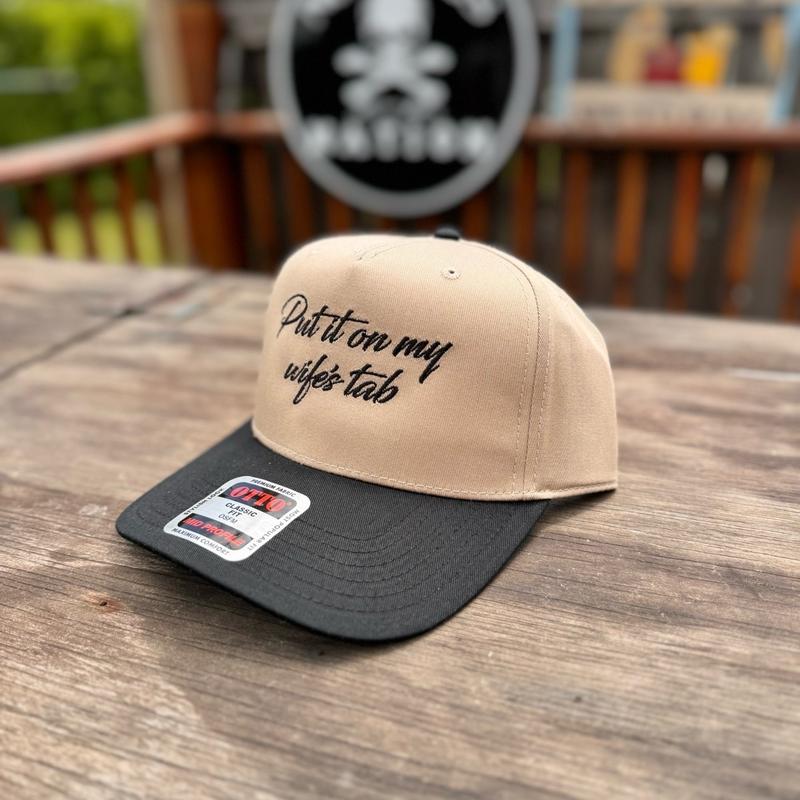 Beige and black cap with text on a wooden surface, brand logo in the background