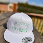 White snapback hat with green text 'Stay Howdy Stay Rowdy' on a wooden surface with blurred background