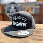Black snapback hat with 'Say Howdy Stay Rowdy' white text on a wooden surface.