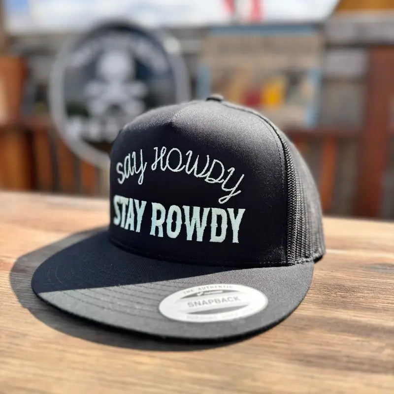 Black snapback hat with 'Say Howdy Stay Rowdy' white text on a wooden surface.