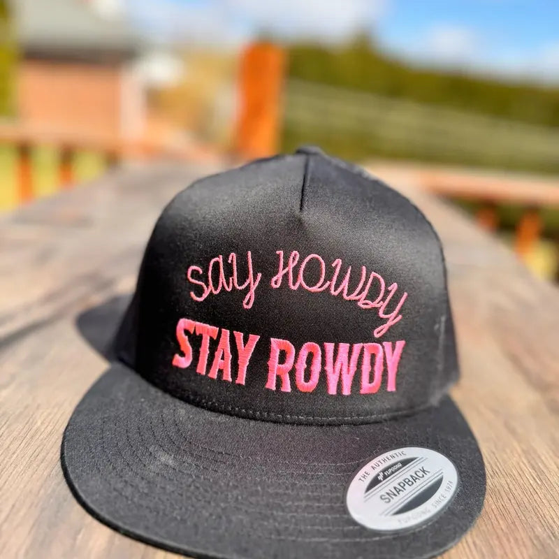 Black snapback hat with pink text 'Say Howdy Stay Rowdy' on a wooden surface