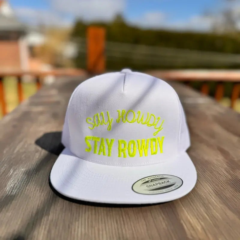White snapback hat with yellow text 'Stay  Howdy Stay Rowdy' on a wooden surface
