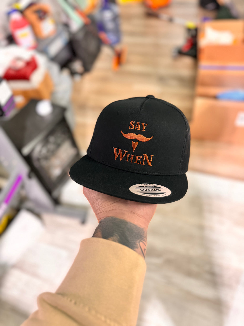 Black snapback hat with orange 'SAY WHEN' text held by a person in a store setting