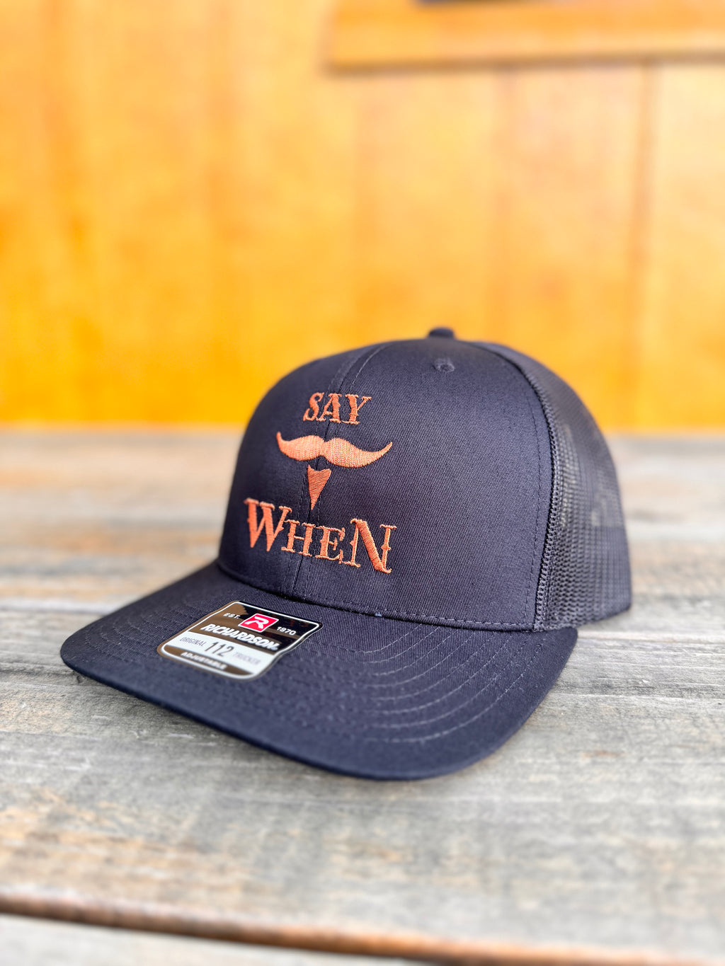Black richardson 112 snapback hat with orange 'SAY WHEN' text on a wooden table