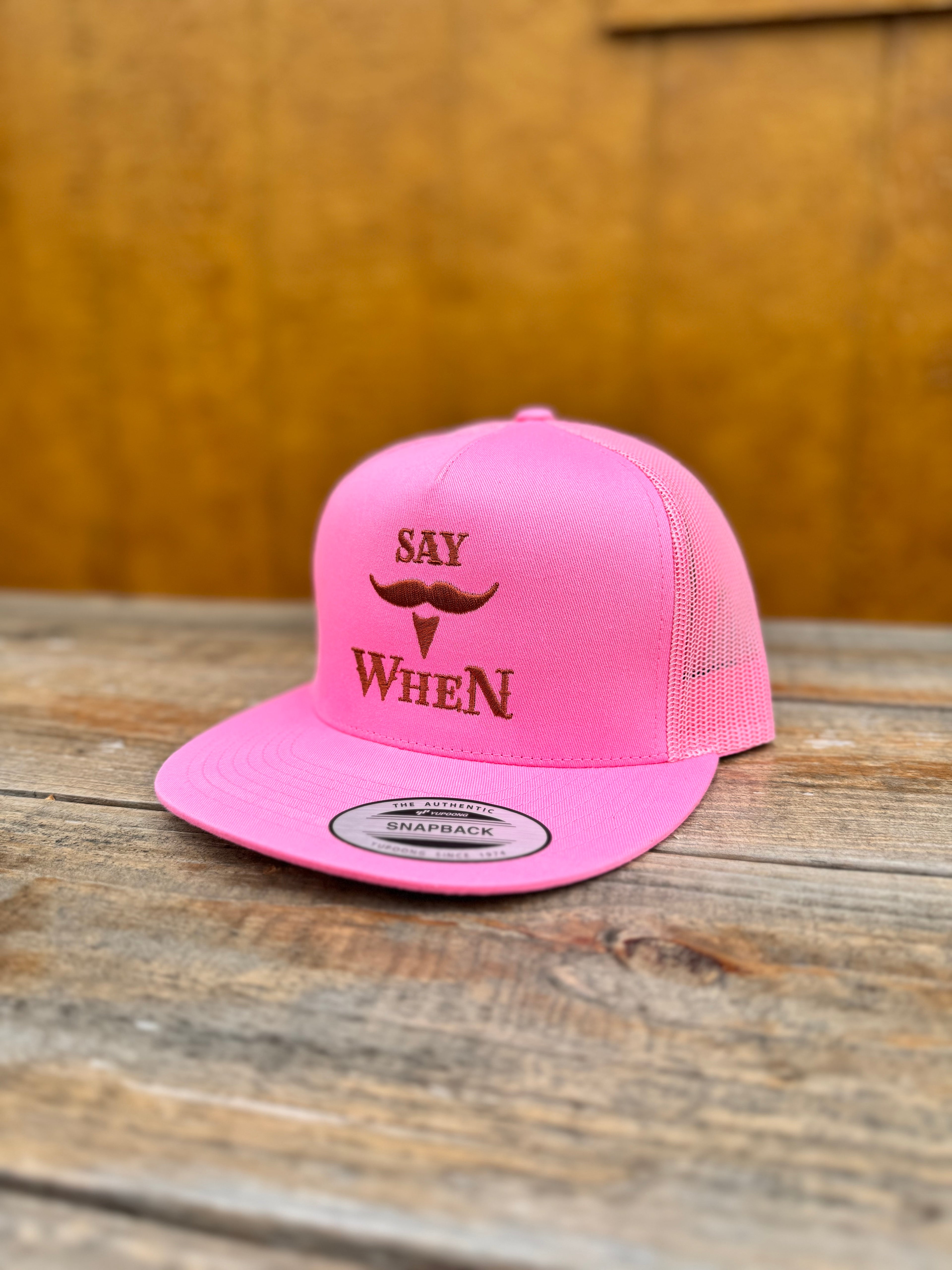 pink snapback hat with orange 'SAY WHEN' text on a wooden table