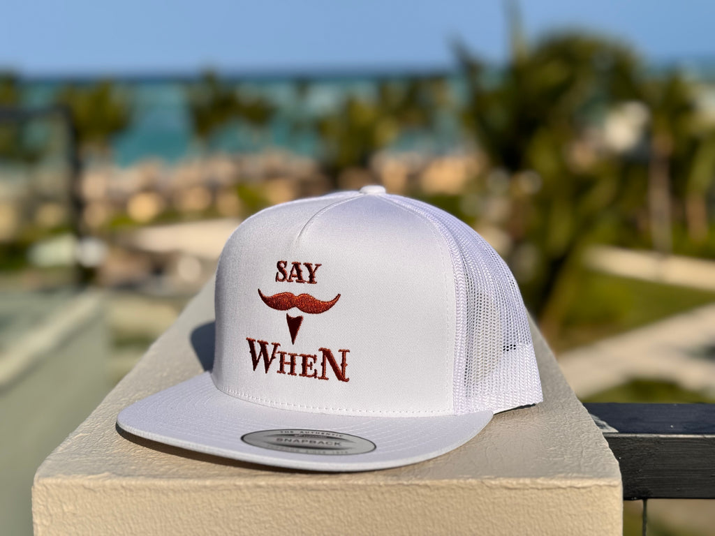 white snapback hat with orange 'SAY WHEN' text on the edge of a balcony in a beachy background