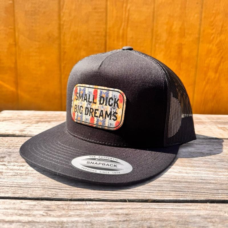 Black trucker cap snapback hat with 'Small D*ck Big Dreams' patch on a wooden surface.