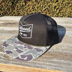 Black trucker snapback hat with camouflage brim and 'Tattooed Low Life' text on a wooden surface.