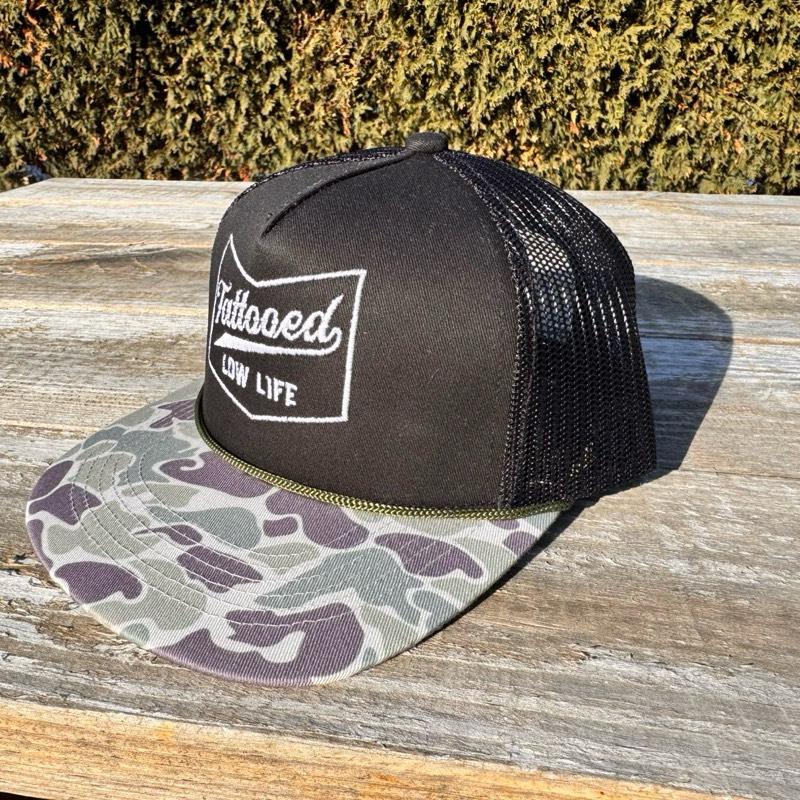 Black trucker snapback hat with camouflage brim and 'Tattooed Low Life' text on a wooden surface.