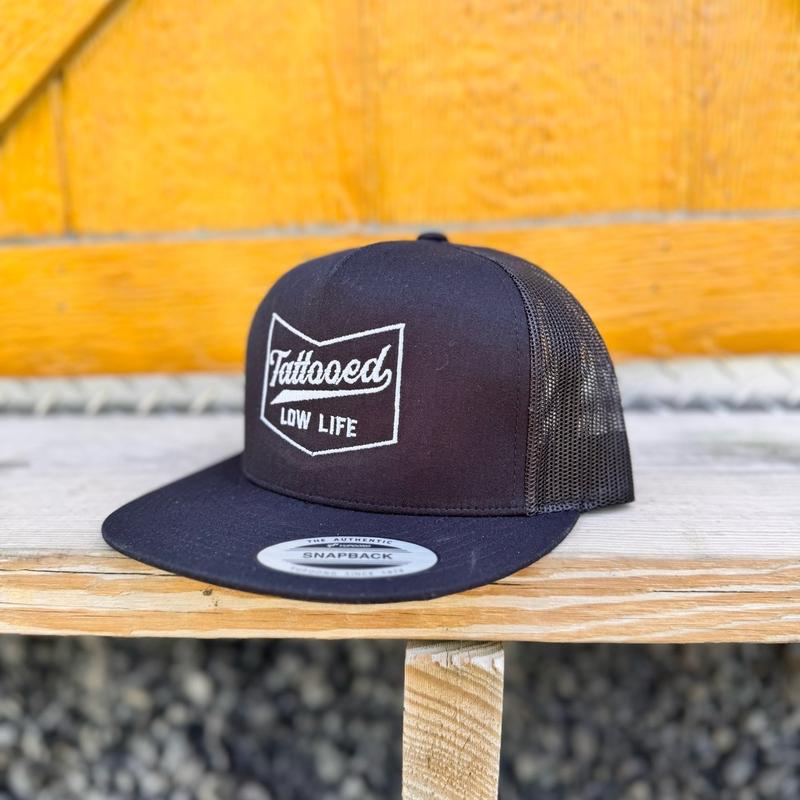 Black snapback hat with 'Tattooed Low Life' logo on a wooden surface.