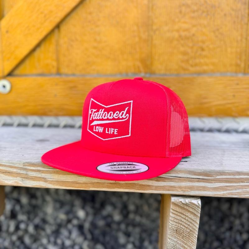 Red snapback hat with white 'tattooed low life' text on a wooden surface