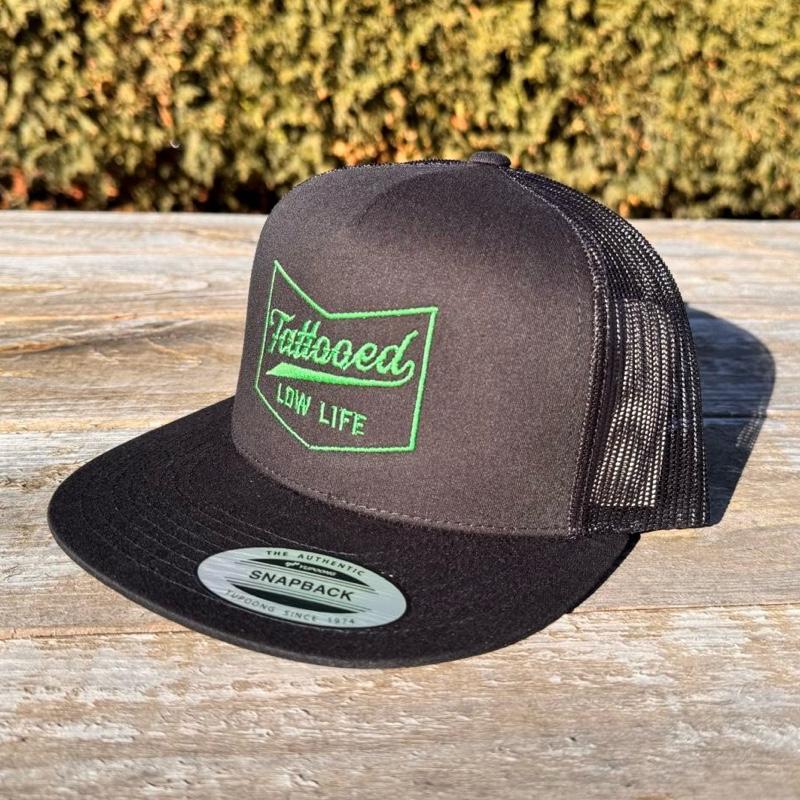 Black snapback hat with green 'Tattooed Low Life' text on a wooden surface with blurred greenery in the background