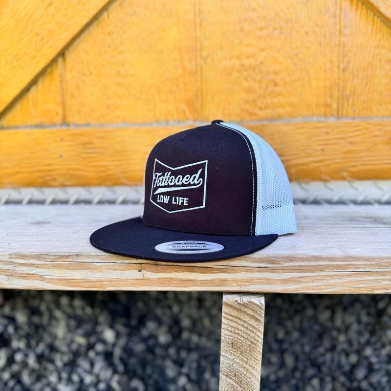 Black and white snapback hat with white 'Tattooed Low Life' text on a wooden surface.
