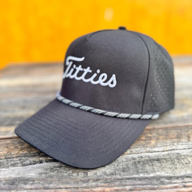 Black snapback hat with 'T*tties' text on a wooden surface with a yellowish background