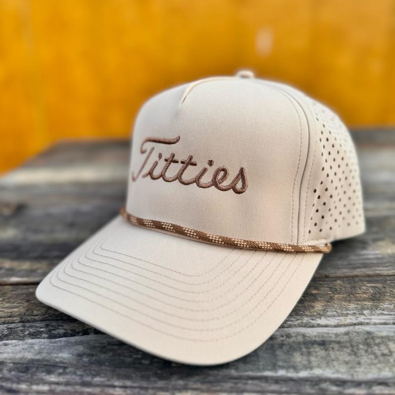 Tan snapback hat with 'T*tties' text on a wooden surface with a yellowish background