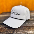 White snapback hat with 'T*tties' text on a wooden surface with a yellowish background