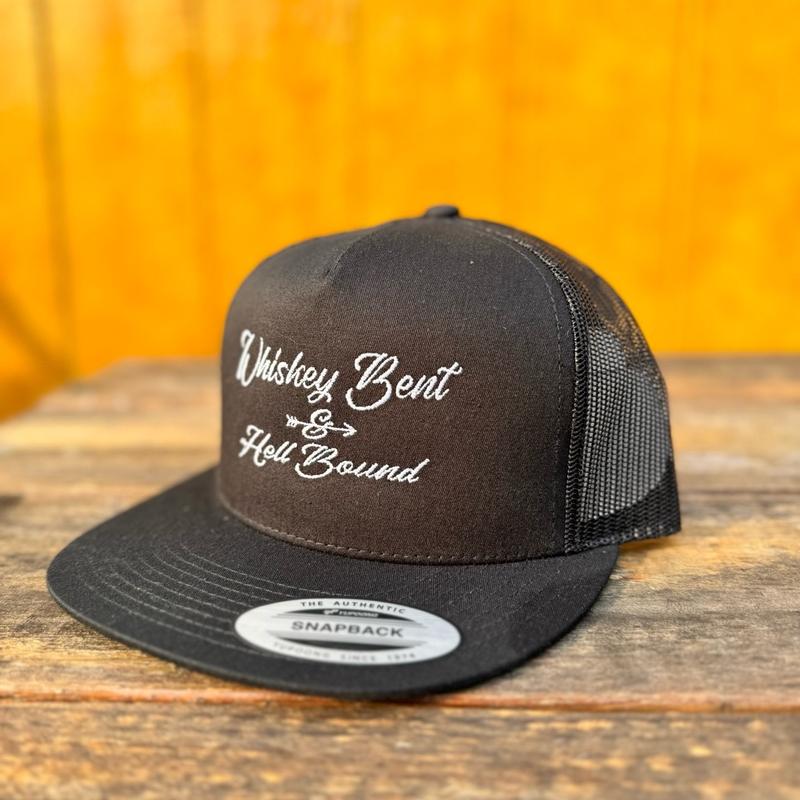 Black trucker snapback hat with 'Whiskey Bent & Hell Bound' text on a wooden surface with orange background